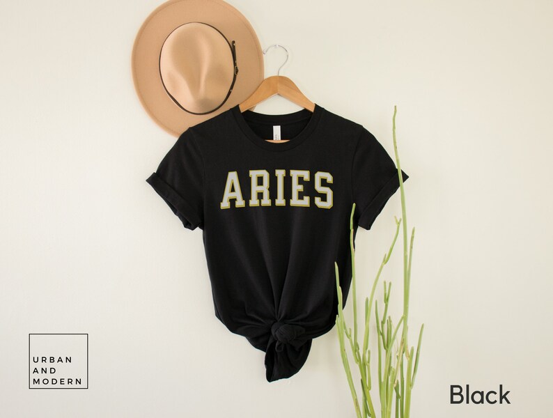 Op de afbeelding: Zwart T-shirt met het woord "ARIES" in een gouden en witte college-lettertype. Het shirt wordt gepresenteerd op een houten hanger met een beige hoed op de achtergrond. De woorden "Urban and Modern" staan in een witte doos linksonder.