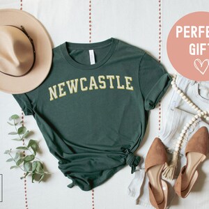 May include: A dark green t-shirt with "NEWCASTLE" in yellow lettering. The shirt is styled with a beige hat, light-wash jeans, and brown flats. A decorative string of beads and a sprig of greenery are also included.