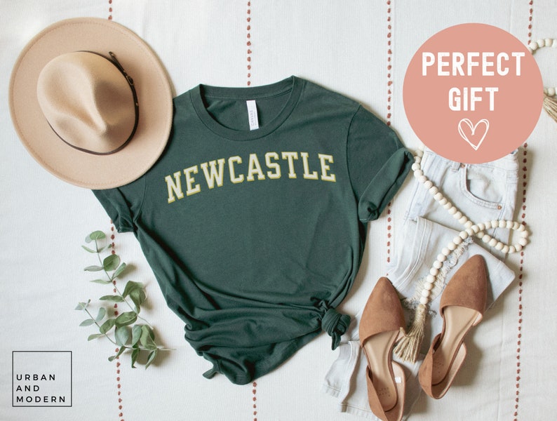 May include: A dark green t-shirt with "NEWCASTLE" in yellow lettering. The shirt is styled with a beige hat, light-wash jeans, and brown flats. A decorative string of beads and a sprig of greenery are also included.