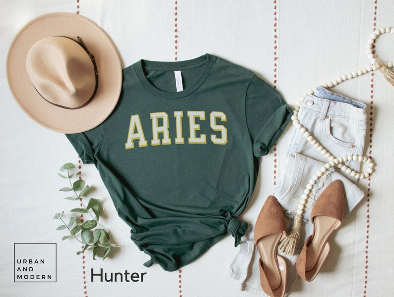 Op de afbeelding: Donkergroen t-shirt met het woord "ARIES" in goud-witte letters. De outfit wordt gecompleteerd door een lichtblauwe jeans, bruine ballerina's en een beige hoed. Een houten kralenketting en een takje groen zijn ook aanwezig.
