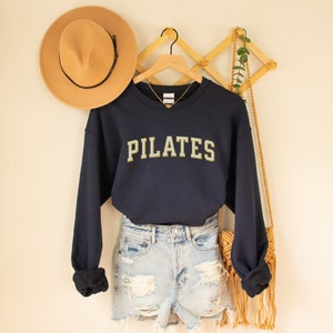 May include: A navy blue sweatshirt with the word "PILATES" in gold lettering. The sweatshirt is worn over a pair of light blue denim shorts and a straw bag.
