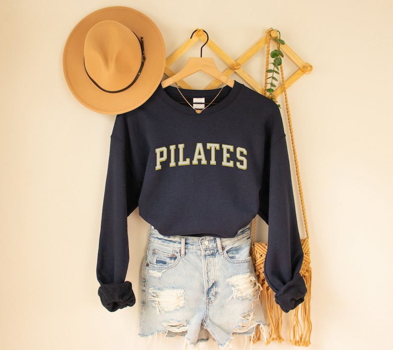 May include: A navy blue sweatshirt with the word "PILATES" in gold lettering. The sweatshirt is worn over a pair of light blue denim shorts and a straw bag.