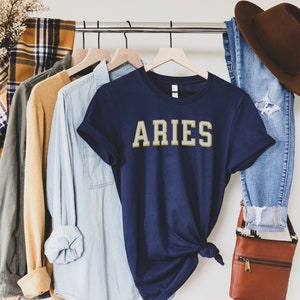 Op de afbeelding: Een marineblauw t-shirt met het woord "ARIES" in gouden letters. De shirt maakt deel uit van een kledingdisplay met een bruine hoed, een blauwe spijkerbroek en andere shirts in verschillende kleuren. De woorden "Urban and Modern" staan in een witte doos linksonder.