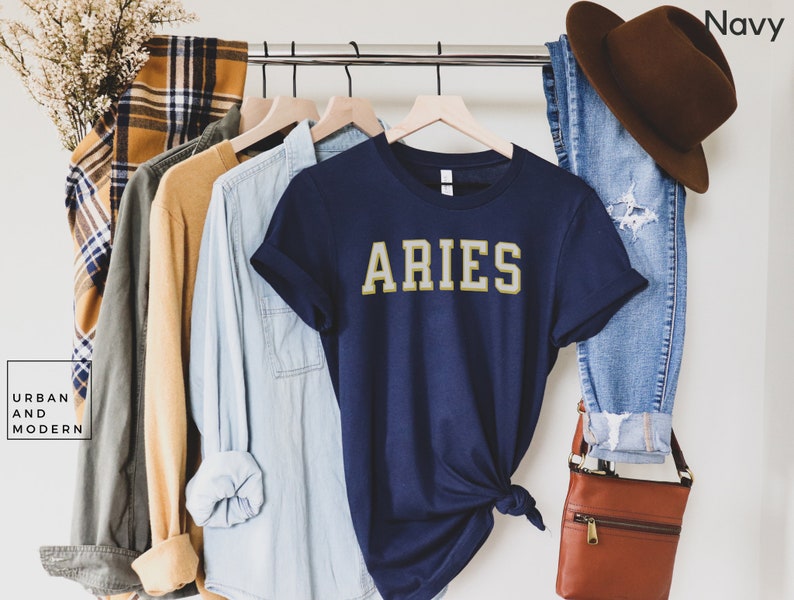 Op de afbeelding: Een marineblauw t-shirt met het woord "ARIES" in gouden letters. De shirt maakt deel uit van een kledingdisplay met een bruine hoed, een blauwe spijkerbroek en andere shirts in verschillende kleuren. De woorden "Urban and Modern" staan in een witte doos linksonder.