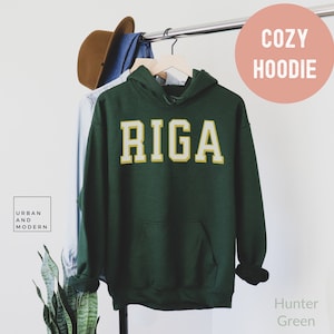 Puede incluir: Sudadera con capucha verde cazador con la palabra "RIGA" en letras blancas y doradas. La sudadera con capucha está colgada en una percha con un sombrero marrón y una camisa vaquera. Las palabras "Urban and Modern" están en un recuadro blanco.