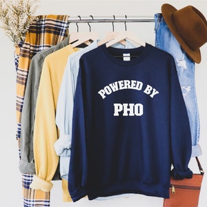 May include: A navy blue sweatshirt with the text "Powered by Pho" in white lettering. The sweatshirt is hanging on a rack with other clothing items.