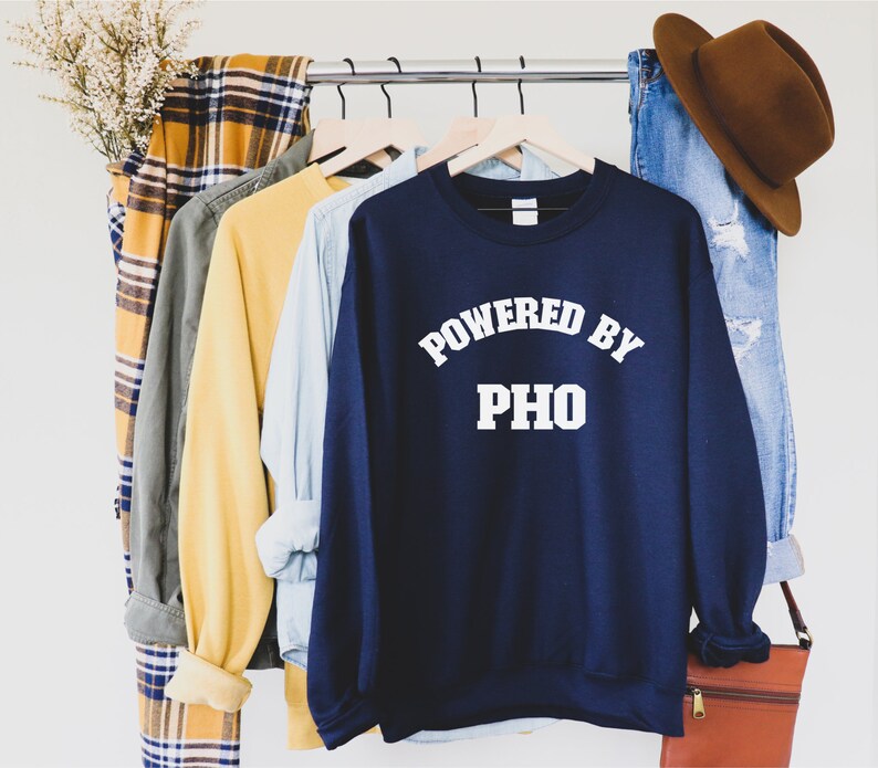 May include: A navy blue sweatshirt with the text "Powered by Pho" in white lettering. The sweatshirt is hanging on a rack with other clothing items.