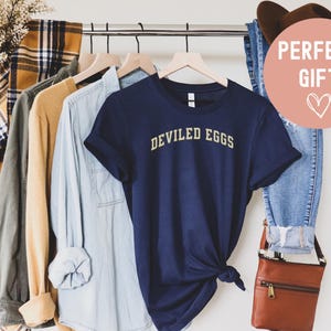 May include: A navy blue t-shirt with the words "DEVILED EGGS" in gold lettering. The shirt is hanging on a wooden hanger with other shirts and a pair of jeans. A brown leather crossbody bag and a brown hat are also visible.