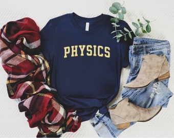 May include: A navy blue t-shirt with the word "PHYSICS" in yellow lettering. The shirt is laid out on a white surface with a plaid blanket and a pair of blue jeans and brown ankle boots.