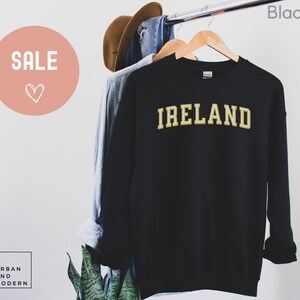 Może przedstawiać: Czarna bluza z napisem "IRELAND" w złotych literach. Bluza wisi na drewnianym wieszaku. Słowo "SALE" znajduje się w r&oacute;żowym okręgu w lewym g&oacute;rnym rogu.