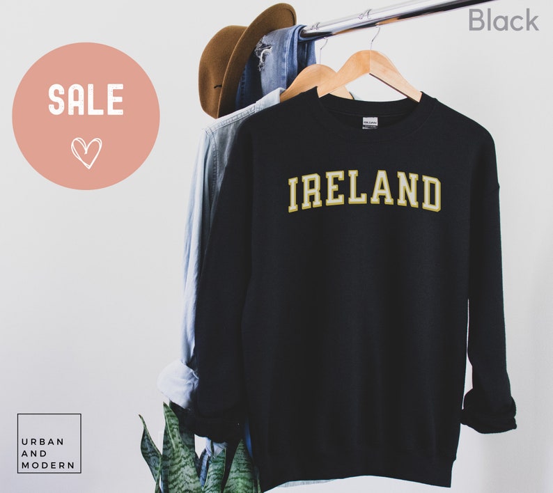 Może przedstawiać: Czarna bluza z napisem "IRELAND" w złotych literach. Bluza wisi na drewnianym wieszaku. Słowo "SALE" znajduje się w r&oacute;żowym okręgu w lewym g&oacute;rnym rogu.