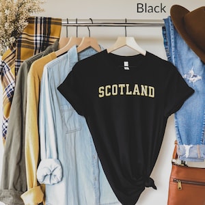 May include: Black t-shirt with the word "SCOTLAND" in gold lettering. The shirt is hanging on a rack with other clothing items.