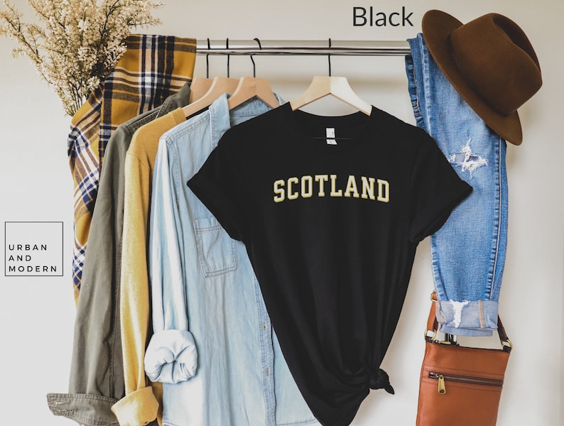 May include: Black t-shirt with the word "SCOTLAND" in gold lettering. The shirt is hanging on a rack with other clothing items.