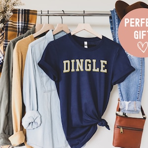 May include: Navy blue t-shirt with the word "DINGLE" in gold lettering. The shirt is hanging on a rack with other clothing items.