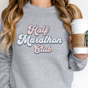 K&ouml;nnte beinhalten: Grauer Pullover mit dem Text "Half Marathon Club" in rosafarbenen und blauen Retro-Lettern.