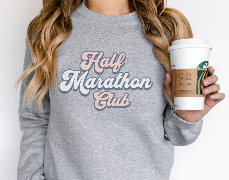 K&ouml;nnte beinhalten: Grauer Pullover mit dem Text "Half Marathon Club" in rosafarbenen und blauen Retro-Lettern.