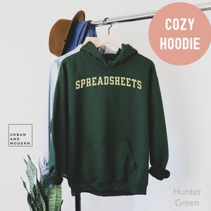May include: A hunter green hoodie with the word "SPREADSHEETS" in yellow lettering. The hoodie is hanging on a hanger and has a pouch pocket.