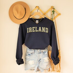 Może przedstawiać: Granatowa bluza z napisem "IRELAND" w złotych literach. Bluza wisi na drewnianym wieszaku, w połączeniu z dżinsowymi szortami i brązowym kapeluszem. Widoczny jest r&oacute;wnież złoty naszyjnik i tkana torba.