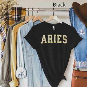 Op de afbeelding: Zwart T-shirt met het woord "ARIES" in gouden letters. Het shirt hangt aan een kledingrek met andere shirts en een spijkerbroek. Het woord "Black" staat boven het shirt. Het merk "URBAN AND MODERN" staat linksonder.
