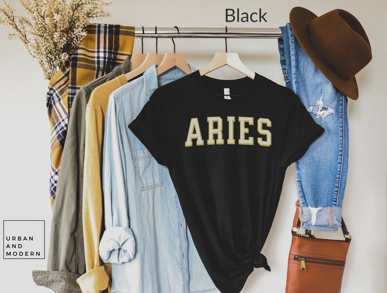 Op de afbeelding: Zwart T-shirt met het woord "ARIES" in gouden letters. Het shirt hangt aan een kledingrek met andere shirts en een spijkerbroek. Het woord "Black" staat boven het shirt. Het merk "URBAN AND MODERN" staat linksonder.