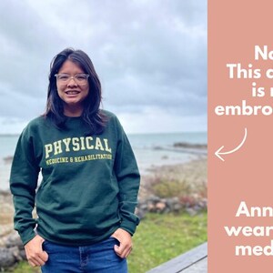 May include: A dark green sweatshirt with the text "PHYSICAL MEDICINE & REHABILITATION" in a gold font. The sweatshirt is worn by a person in front of a coastal background. Text on the side says "Note This design is not embroidered."