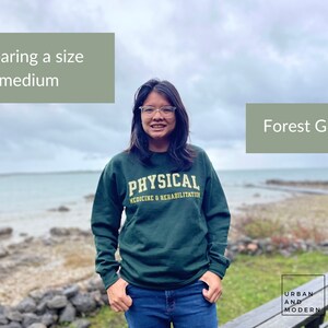 Pu&ograve; includere: Una donna indossa una felpa verde bosco con il testo "PHYSICAL MEDICINE & REHABILITATION" in giallo. &Egrave; in piedi di fronte a un specchio d'acqua e una costa rocciosa. Il testo "Wearing a size medium" e "Forest Green" si trova negli angoli superiori dell'immagine.