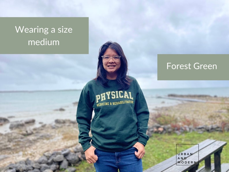 Pu&ograve; includere: Una donna indossa una felpa verde bosco con il testo "PHYSICAL MEDICINE & REHABILITATION" in giallo. &Egrave; in piedi di fronte a un specchio d'acqua e una costa rocciosa. Il testo "Wearing a size medium" e "Forest Green" si trova negli angoli superiori dell'immagine.