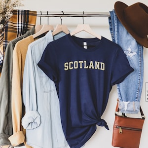 May include: A navy blue t-shirt with the word "SCOTLAND" in yellow lettering. The shirt is hanging on a rack with other clothing items.
