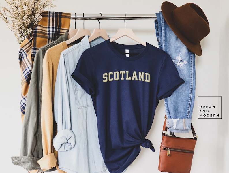 May include: A navy blue t-shirt with the word "SCOTLAND" in yellow lettering. The shirt is hanging on a rack with other clothing items.