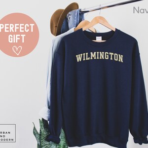 May include: Navy blue sweatshirt with "WILMINGTON" in gold lettering. The sweatshirt is hanging on a wooden hanger with a light blue shirt and a brown hat. The words "PERFECT GIFT" are in a pink circle.