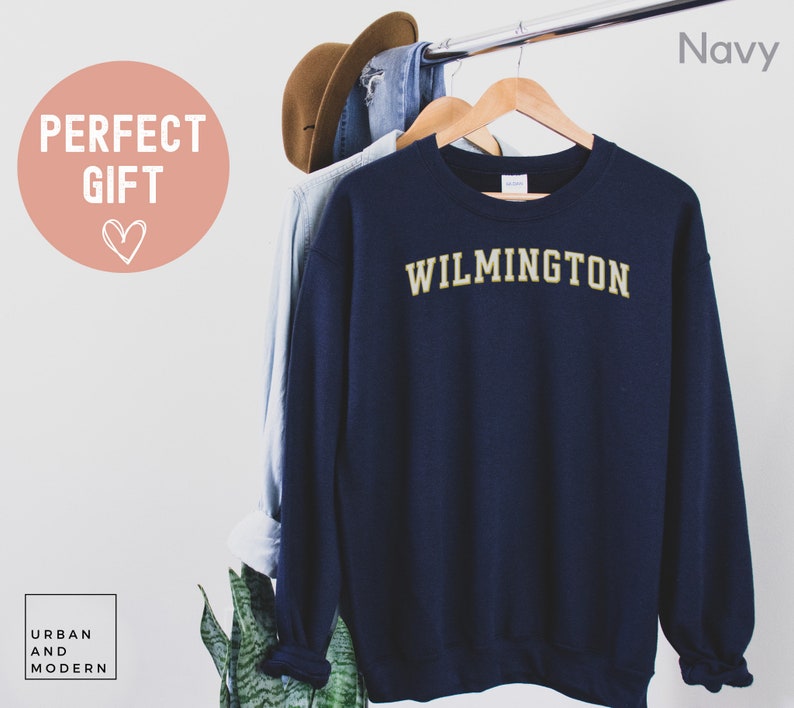 May include: Navy blue sweatshirt with "WILMINGTON" in gold lettering. The sweatshirt is hanging on a wooden hanger with a light blue shirt and a brown hat. The words "PERFECT GIFT" are in a pink circle.