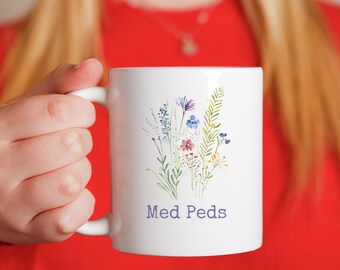 Könnte beinhalten: Weiße Keramiktasse mit einem Aquarell-Blumenmuster in Lila, Blau, Rot und Grün. Die Tasse trägt den Text "Med Peds" in blauer Schrift. Die Tasse wird von einer Person gehalten, die ein rotes Oberteil trägt.