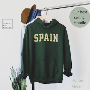 May include: A hunter green hoodie with a gold "SPAIN" graphic on the front. The hoodie is hanging on a hanger.