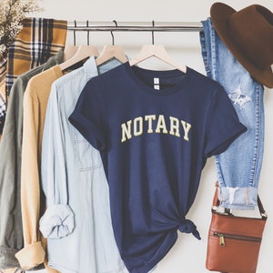 May include: A navy blue t-shirt with the word "NOTARY" in yellow letters. The shirt is hanging on a rack with other clothing items, including a plaid shirt, a tan sweater, and a light blue denim shirt.