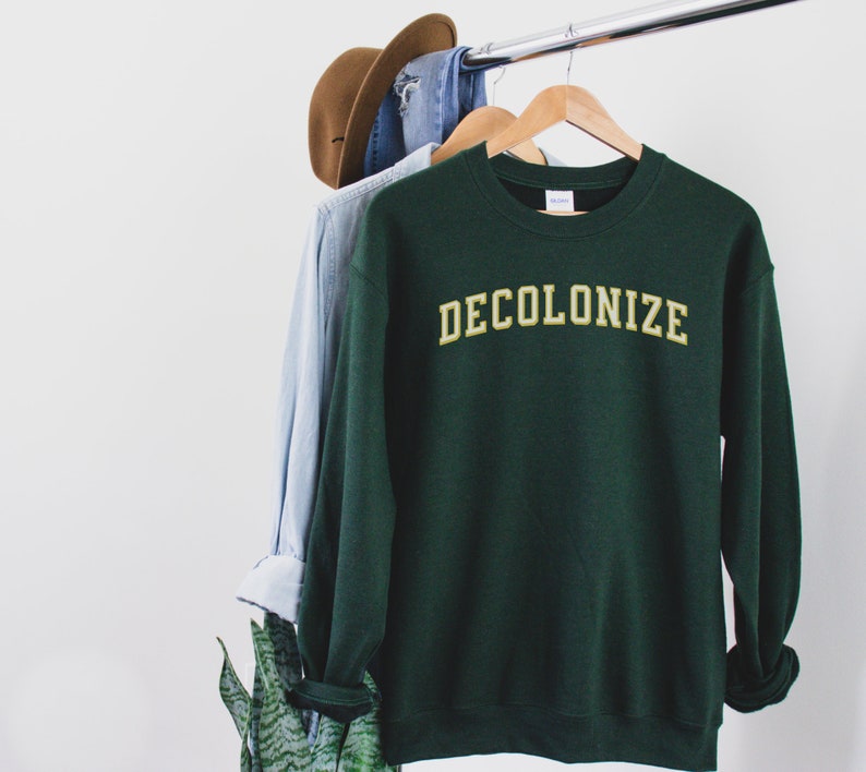 Pu&ograve; includere: Una felpa verde scuro con la scritta "DECOLONIZE" in lettere gialle sul davanti.