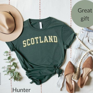 May include: A dark green t-shirt with the word "SCOTLAND" in yellow letters. The shirt is laid out on a white surface with a hat, jeans, and shoes.