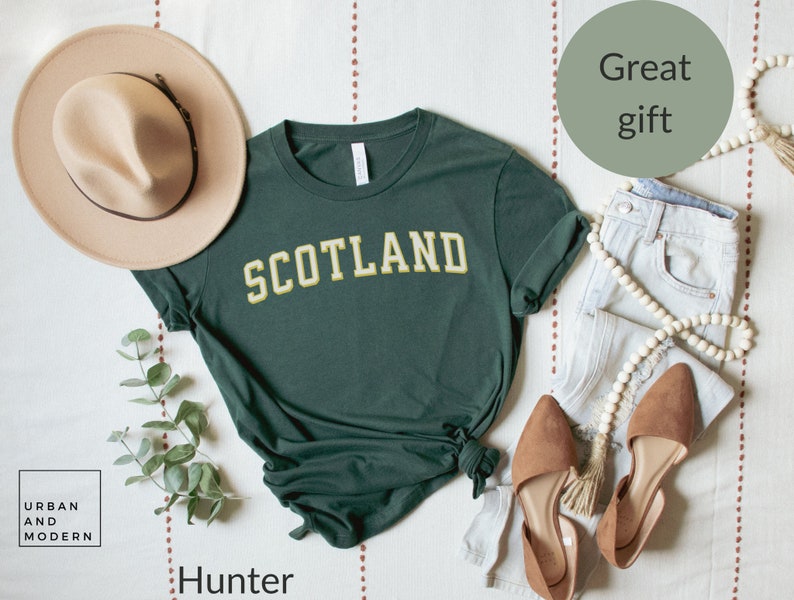 May include: A dark green t-shirt with the word "SCOTLAND" in yellow letters. The shirt is laid out on a white surface with a hat, jeans, and shoes.