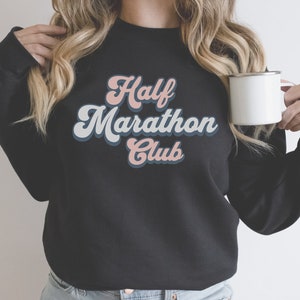 K&ouml;nnte beinhalten: Schwarzes Sweatshirt mit einer Retro-Grafik, auf der "Half Marathon Club" in rosa und blauen Buchstaben steht.
