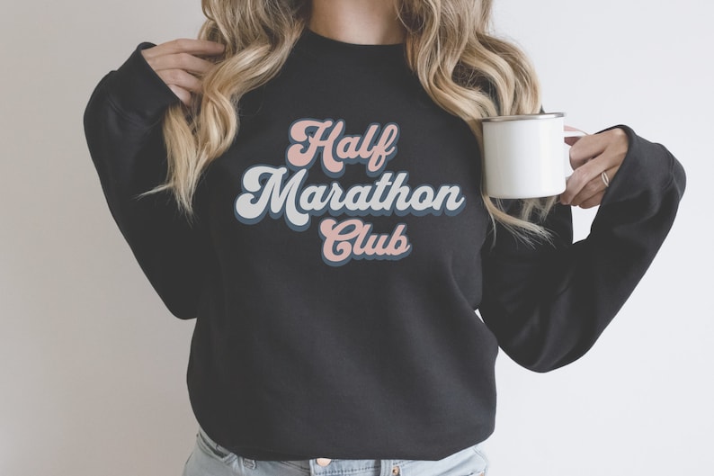 K&ouml;nnte beinhalten: Schwarzes Sweatshirt mit einer Retro-Grafik, auf der "Half Marathon Club" in rosa und blauen Buchstaben steht.