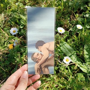 Könnte beinhalten: Ein rechteckiger Lesezeichen mit einer Illustration von zwei Männern, die sich umarmen. Das Kunstwerk verwendet eine weiche Farbpalette. Das Lesezeichen wird vor einem Hintergrund aus grünem Gras und weißen Gänseblümchen gehalten.