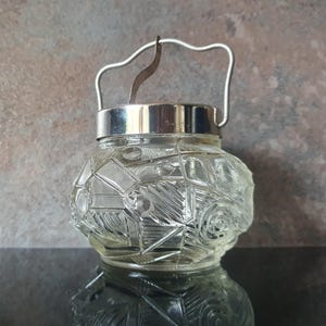 May include: A clear glass jar with a silver rim and handle. The jar features a geometric pattern and a rounded shape. The handle is arched and attached to the rim. The jar is on a reflective surface.