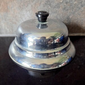 May include: A polished, silver-toned bell with a black top. The bell features a tiered design and reflects its surroundings. This bell is likely used for service or signalling.