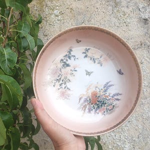 Op de afbeelding: Een ronde, lichtroze keramische kom met een bloemen- en vlinderontwerp. De kom heeft een decoratieve rand en een centraal beeld van bloemen en vlinders in roze, witte en paarse tinten.