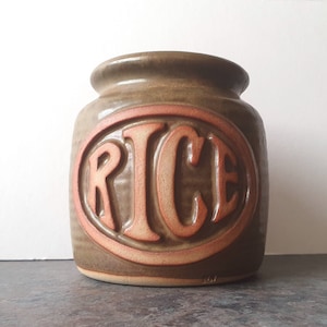 May include: A brown ceramic jar with a red oval label that says "RICE" in raised letters.
