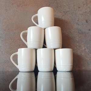 May include: A stack of six white ceramic mugs with rounded handles. The mugs are arranged in a pyramid shape.