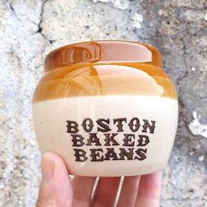 May include: A small, vintage ceramic jar with a brown lid and a cream-coloured body. The words "BOSTON BAKED BEANS" are printed in dark brown, stylised letters on the front. The jar is held in a hand.