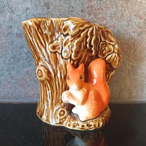 May include: A ceramic squirrel planter shaped like a tree trunk with brown and orange glaze. The squirrel is perched on the trunk with its tail curled around the tree.