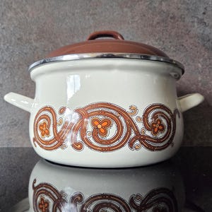 May include: Cream-coloured enamel pot with a brown lid and handle. The pot features a decorative brown and orange floral pattern around the base. It has two side handles and a silver rim.