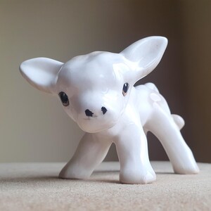 May include: A white ceramic figurine of a calf with black eyes and a brown nose. The calf is standing on all four legs and has its head tilted to the side.