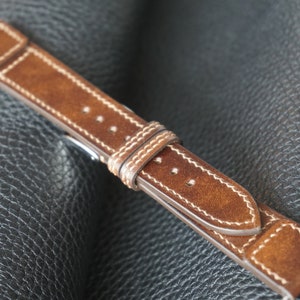 Könnte beinhalten: Ein braunes Lederarmband mit weißen Nähten. Das Armband liegt auf einem schwarzen Lederuntergrund.